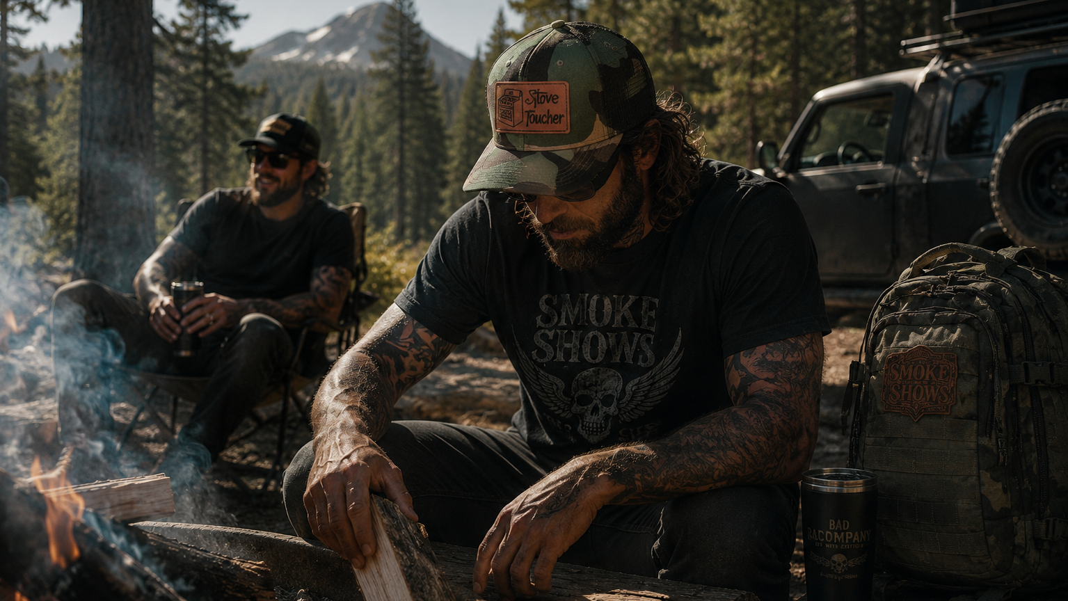 A man in a forest camping setting wearing a black graphic t-shirt with 'SMOKE SHOW' printed on the front, a camouflage cap, and sitting by a campfire next to a rugged off-road vehicle.