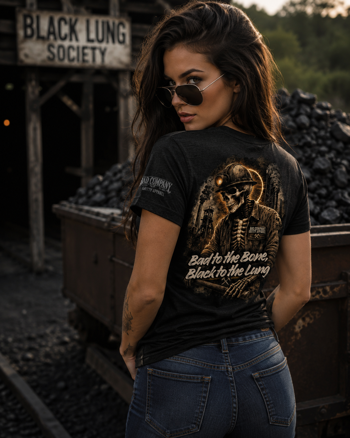 A woman wearing a black graphic t-shirt with a biker skeleton design and the text 'Bad to the Bone, Black to the Lung' on the back, standing in front of a train yard with coal bins.