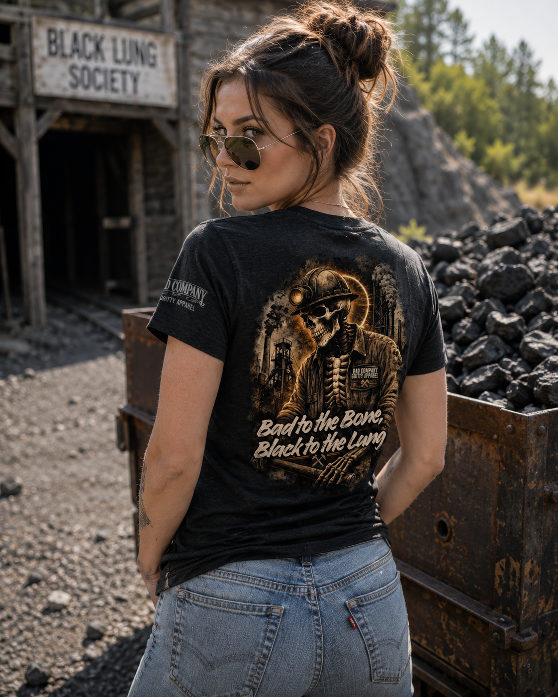 A woman wearing a black graphic t-shirt and light blue jeans stands in a dusty, industrial outdoor setting with a sign reading 'BLACK LUNG SOCIETY' in the background. The back of the t-shirt features a detailed graphic of a skeleton worker in a miner's helmet with the text 'Bad to the Bone.