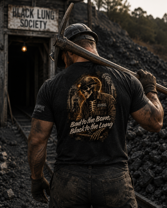 A man viewed from behind wearing a black graphic t-shirt and a hard hat, holding a pickaxe over his shoulder in a dark, gritty outdoor setting with a sign reading 'BLACK LUNG SOCIETY'. The back of the t-shirt features a detailed graphic of a skeleton miner with the text 'Bad to the Bone, Black to the Lung'.
