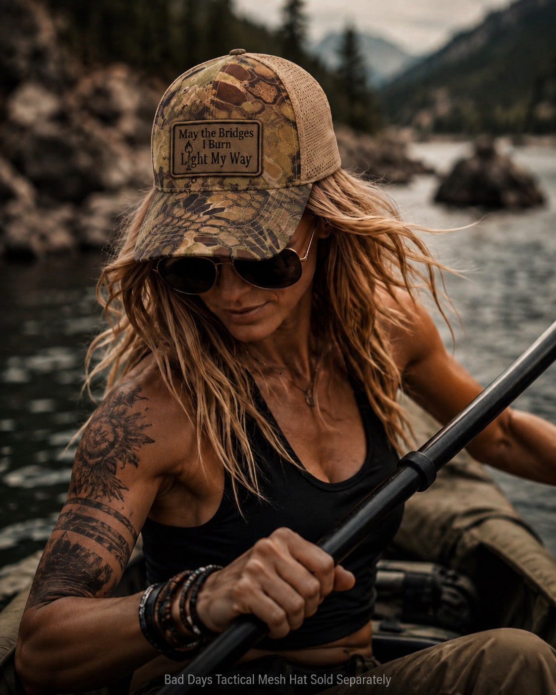 A woman paddling on a river wearing a camouflage trucker hat with a tan front patch that reads 'May the Bridges  I Burn Light My Way' and black sunglasses.