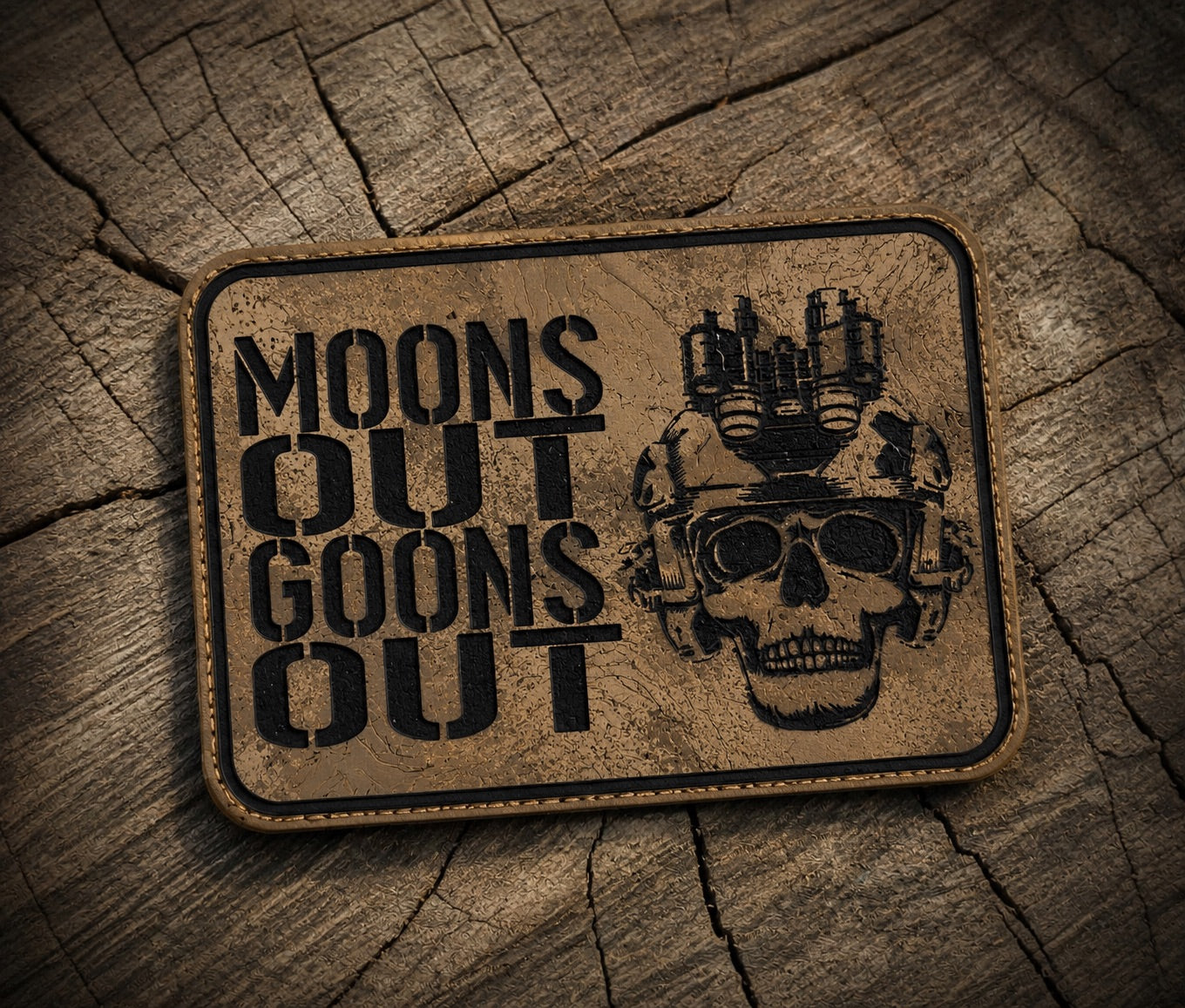 A rectangular tan tactical patch with rounded corners, featuring the text 'MOONS OUT GOONS OUT' in black stencil font alongside a black skull wearing a helmet with mounted lights, placed on a textured wood background.