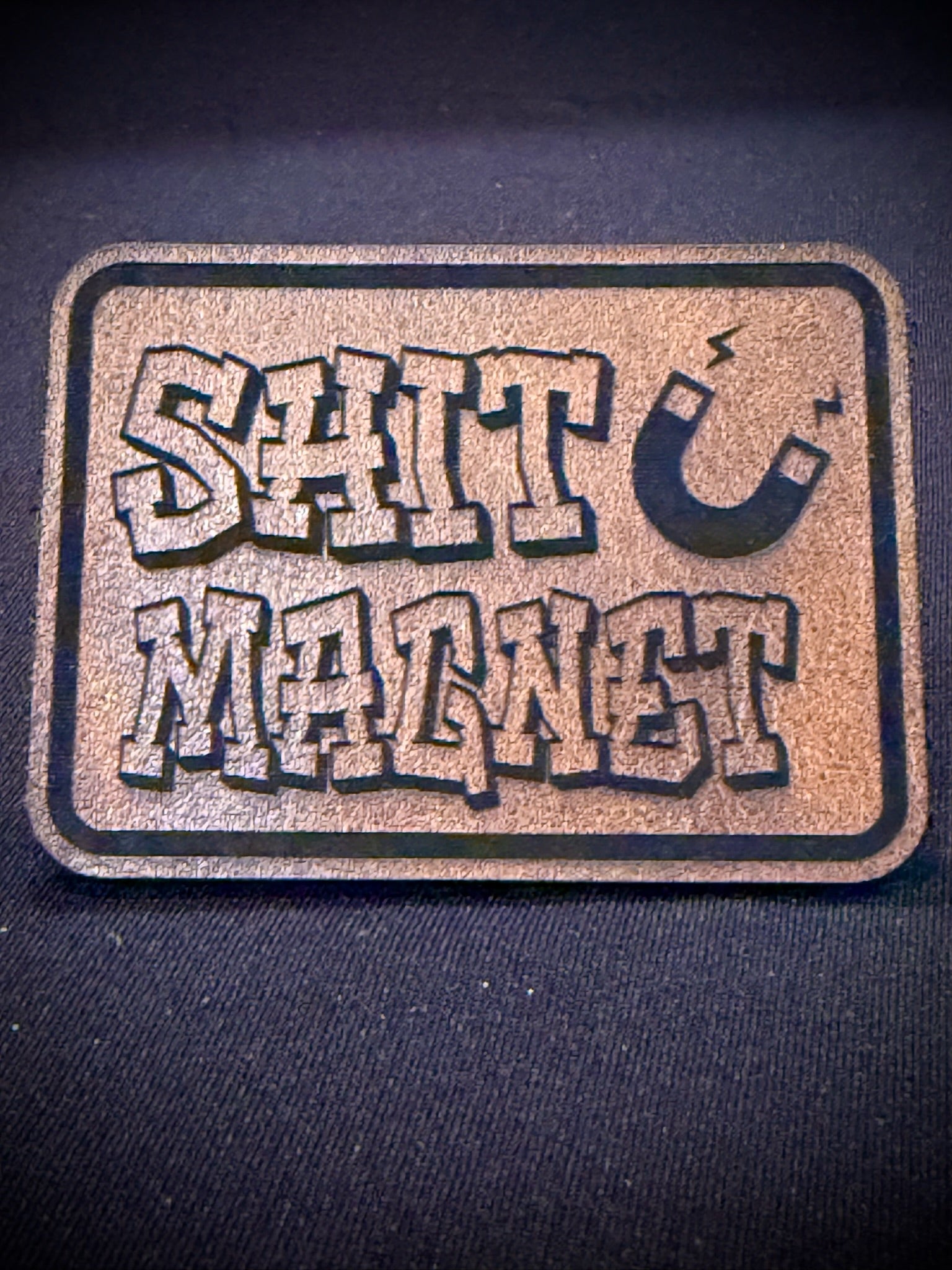 A rectangular morale patch with rounded corners featuring the words 'SHIT MAGNET' in a bold, stylized font and a horseshoe graphic, shown on a dark fabric background.
