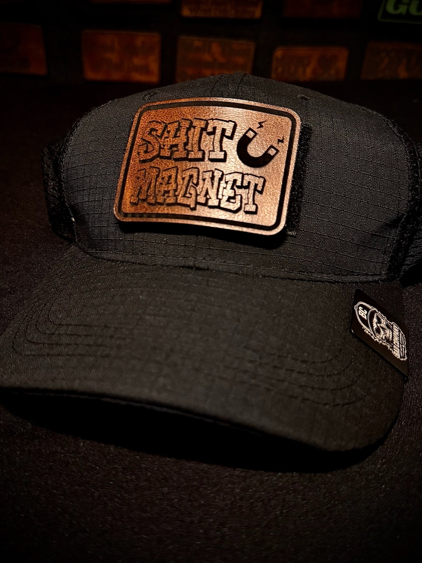 A black Bad Days Tactical Mesh trucker-style cap with a large rectangular front morale patch reading “SHIT MAGNET” in raised lettering, with a small white logo patch on the brim, photographed against a dark background.
