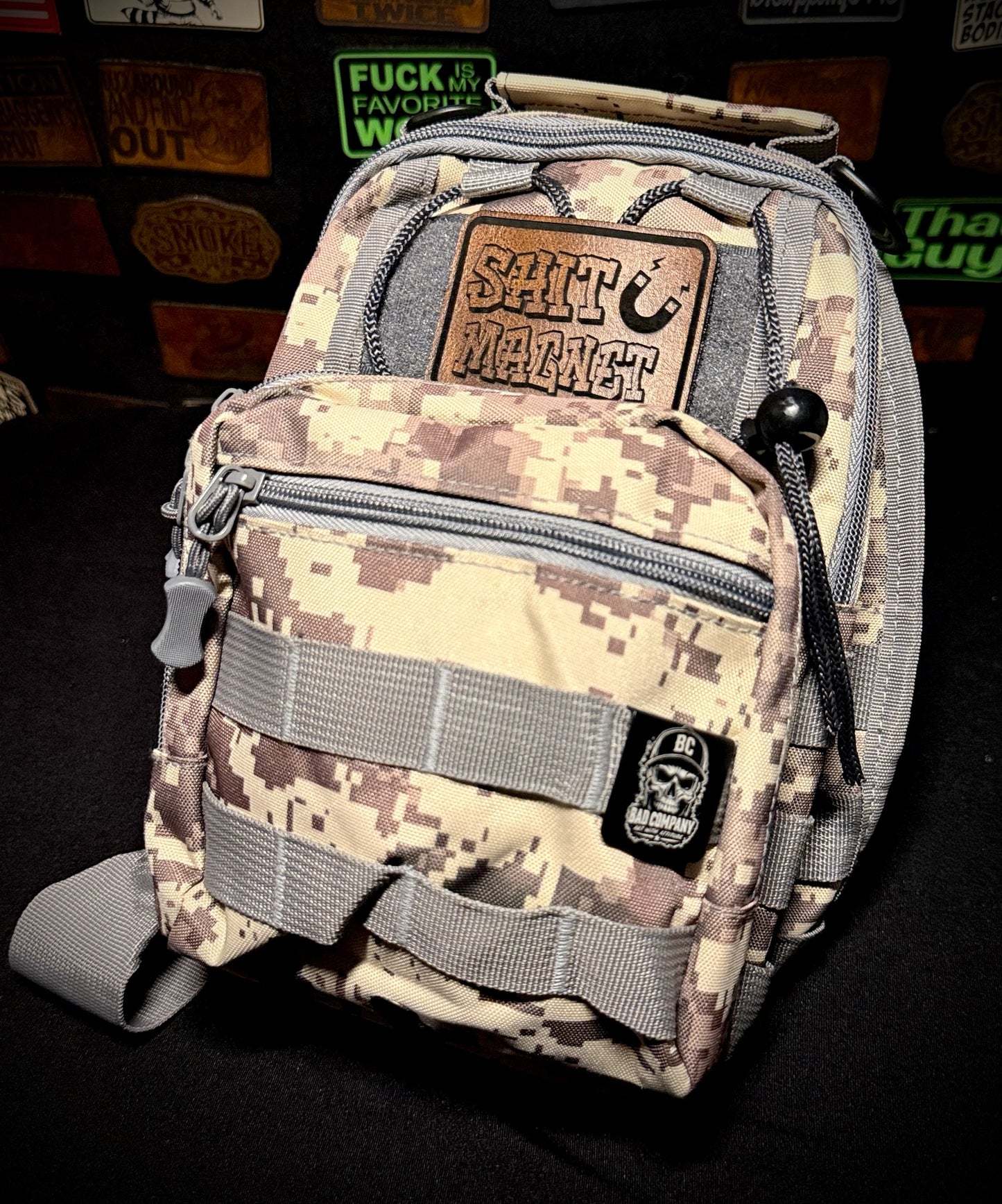 A Bad Decisions Tactical Sling Bag in digital camouflage with gray webbing and zippers, featuring a brown leather 'Shit Magnet' Morale patch on the front pocket and a black skull metal badge below it, set against a dark background with blurred MORALE PATCHES.