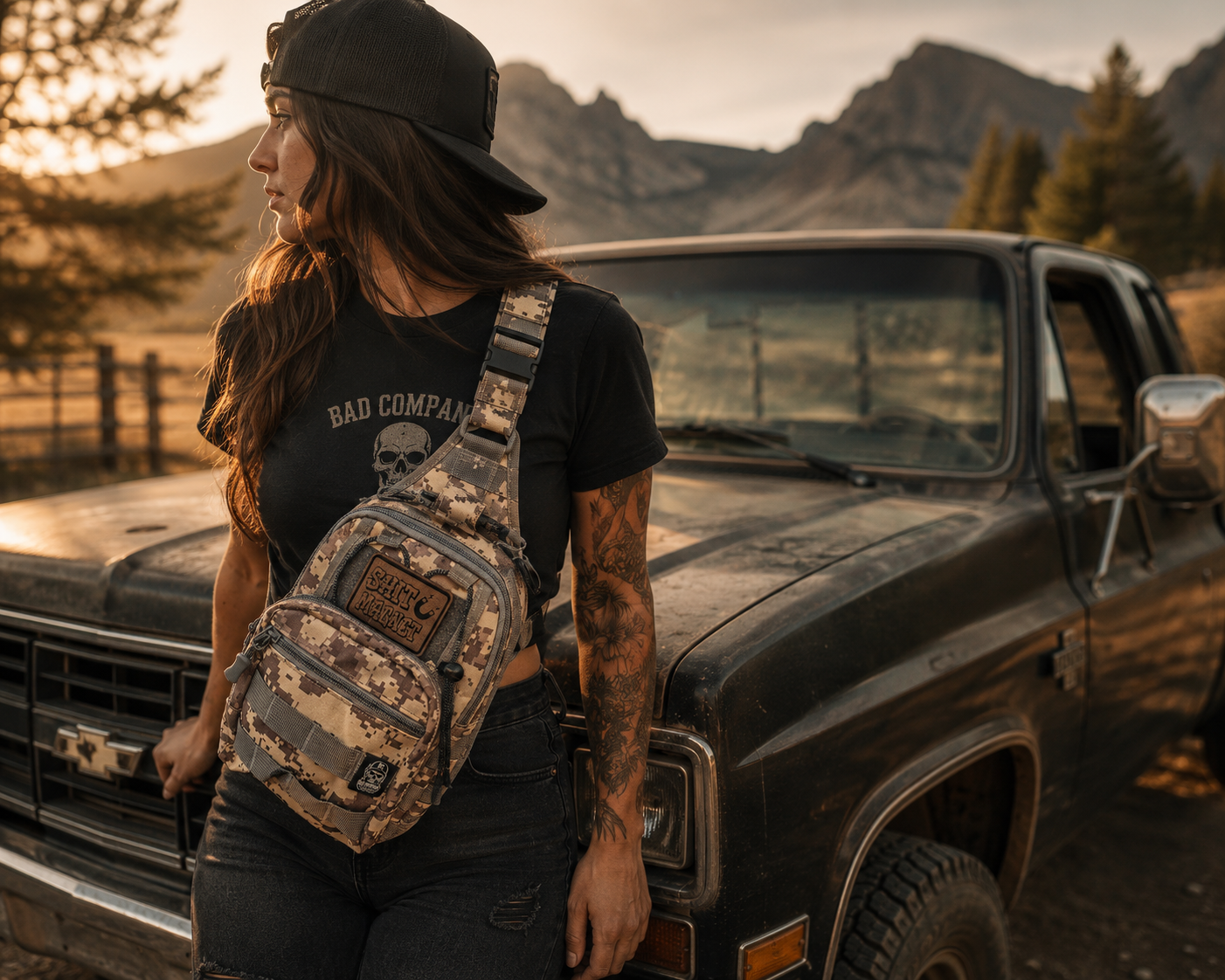 A woman wearing a black cap and a black graphic t-shirt stands next to a vintage pickup truck outdoors, wearing a camouflage patterned Bad Decisions Tactical crossbody sling bag with a brown leather morale patch that reads "Shit Magnet" . The background features a rustic mountain landscape at sunset.