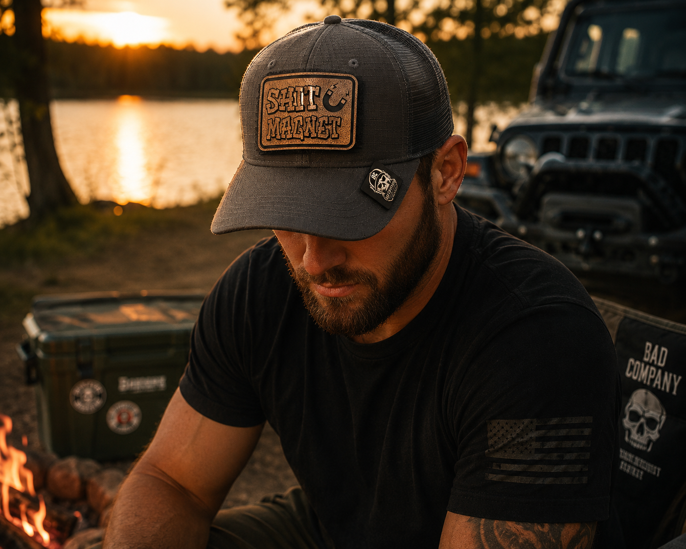 A man wearing a Bad Day Tactical Mesh cap with a tan leather morale patch that reads 'SHIT MAGNET' and a black t-shirt with a skull graphic, sitting outdoors near a campfire and a vehicle at sunset.