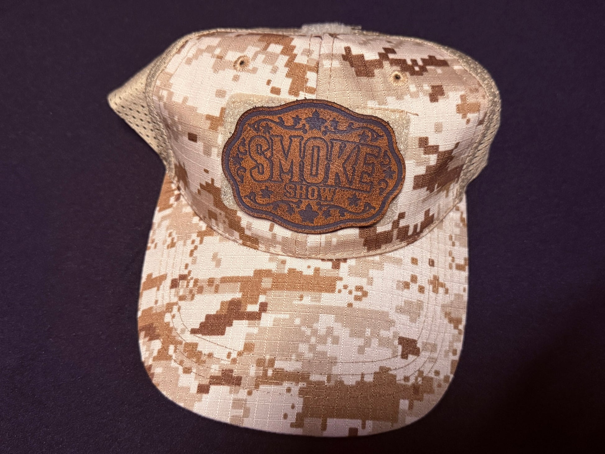 A desert digital camouflage trucker hat with a brown embroidered 'Smoke Show' morale patch on the front, displayed on a dark background.
