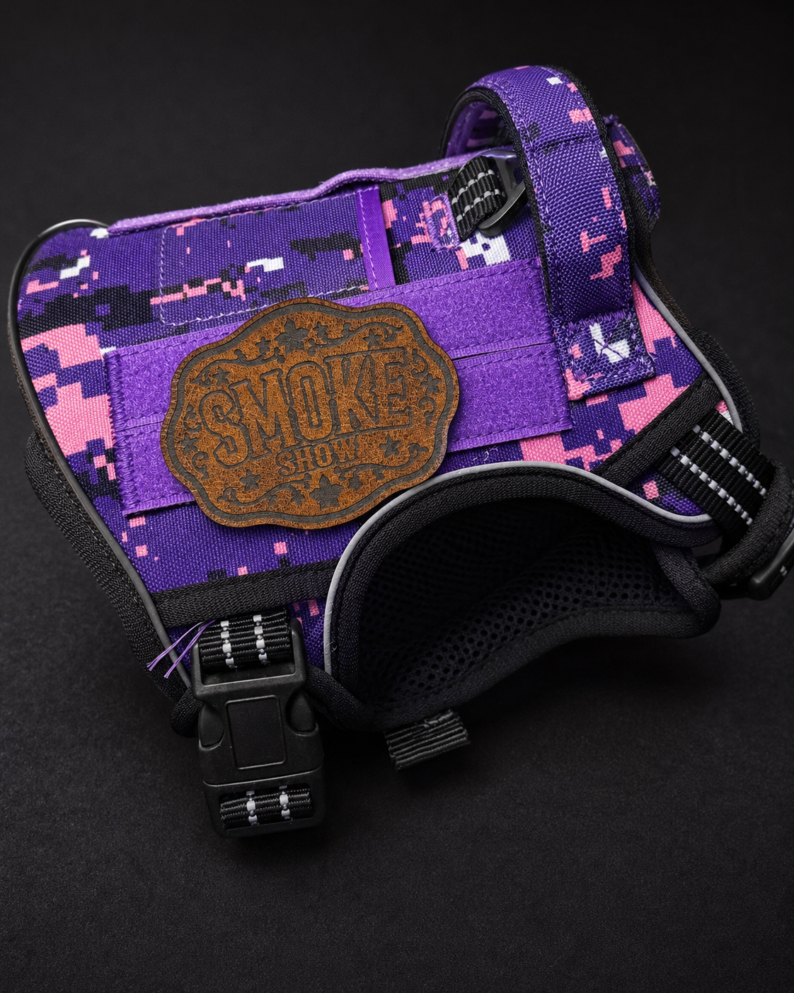 A purple and pink digital camouflage dog harness with a brown leather morale patch reading 'Smoke Show' on the front, shown on a dark background.