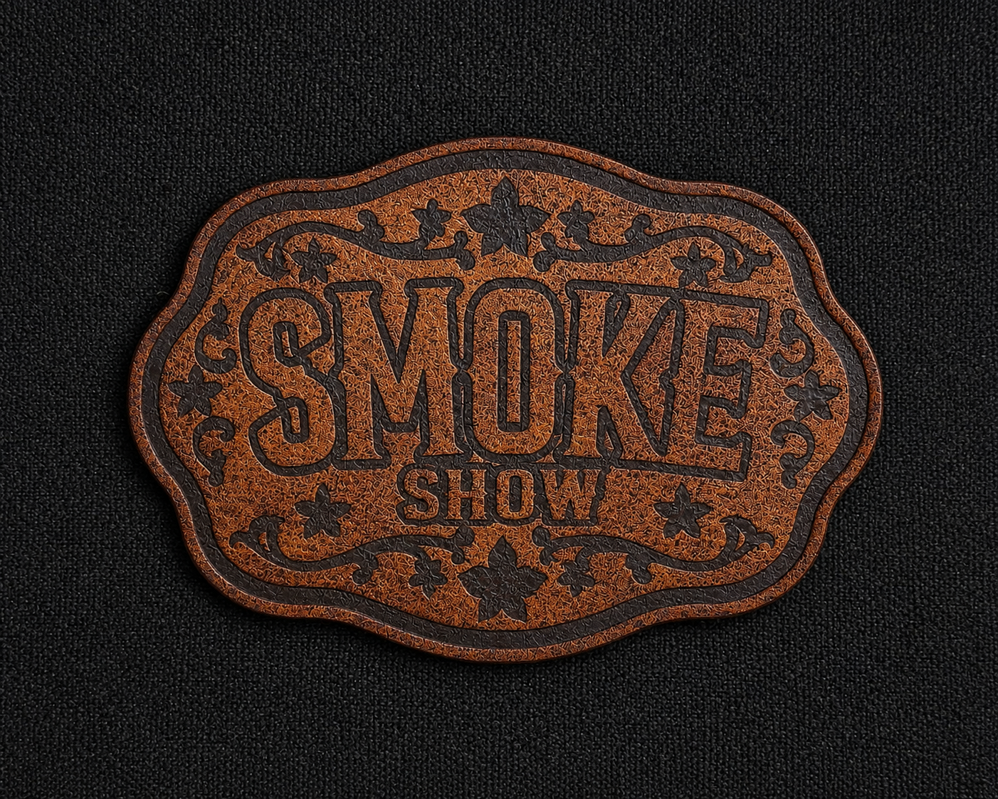 A brown textured leather morale patch with scalloped edges and the words 'SMOKE SHOW' in raised lettering, displayed on a black fabric background.