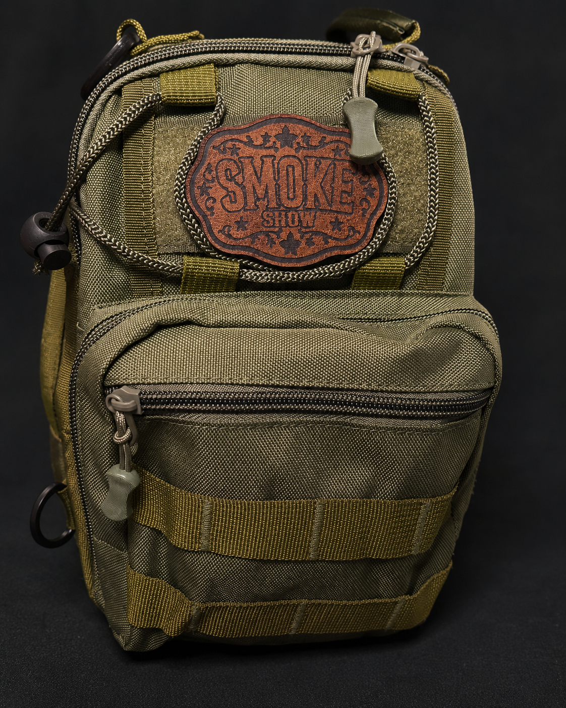 An olive green tactical EC Sling bag with a brown leather 'Smoke Show' morale patch on the front pocket, set against a dark background.