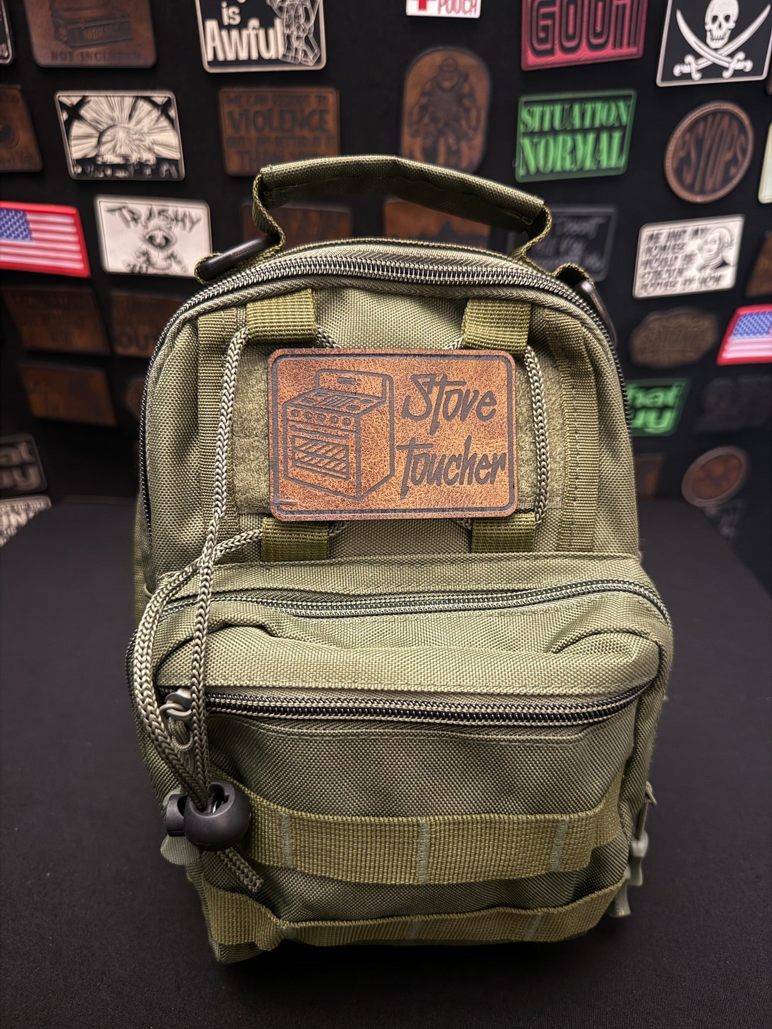 An olive green tactical backpack with a brown leather patch on the front that reads 'Stove Toucher'. The backpack is displayed against a black background covered with various funny morale patches.