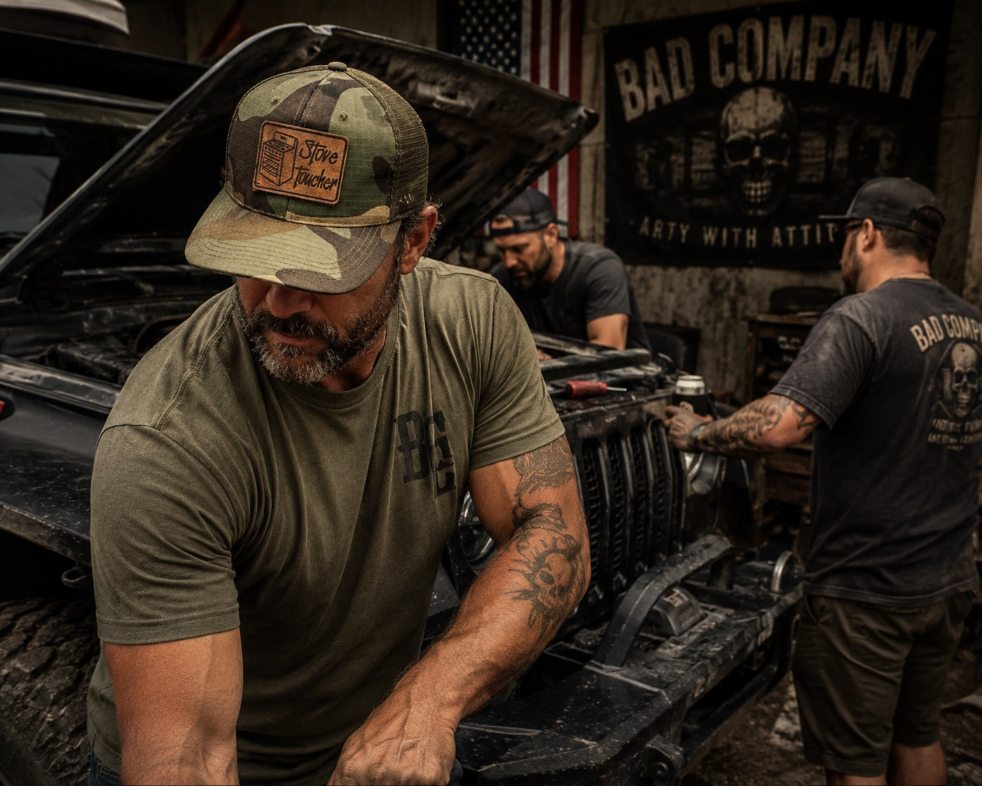 A man in a camouflage truck cap WITH A  FUNNY MORALE PATCH THAT SAYS "STOVE TOUCHER" and olive green t-shirt works on a vehicle in a garage, with another man in a black 'Bad Company' t-shirt and another man in the background near an American flag and a 'Bad Company' banner.